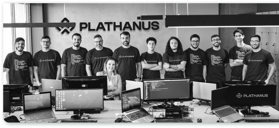 11 people were positioned standing and 1 sitting, all looking forward and wearing the company's uniform. 13 connected monitors are on top of a table demonstrating development code and illustrations. In the background, a sign with the company symbol and written “Plathanus”